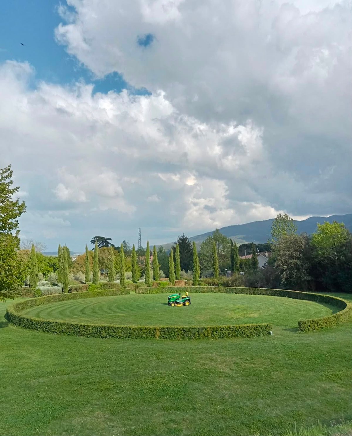 Person on green riding mower in center of large manicured lawn surrounded by cypress trees and mountains under blue cloudy sky